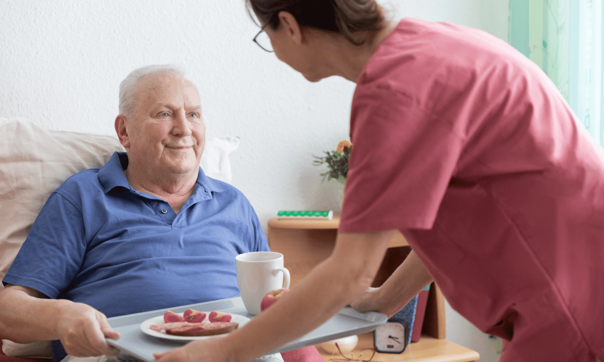 Un cuidador interno atento sirve el desayuno a un hombre mayor sonriente, asegurando su bienestar en el hogar.