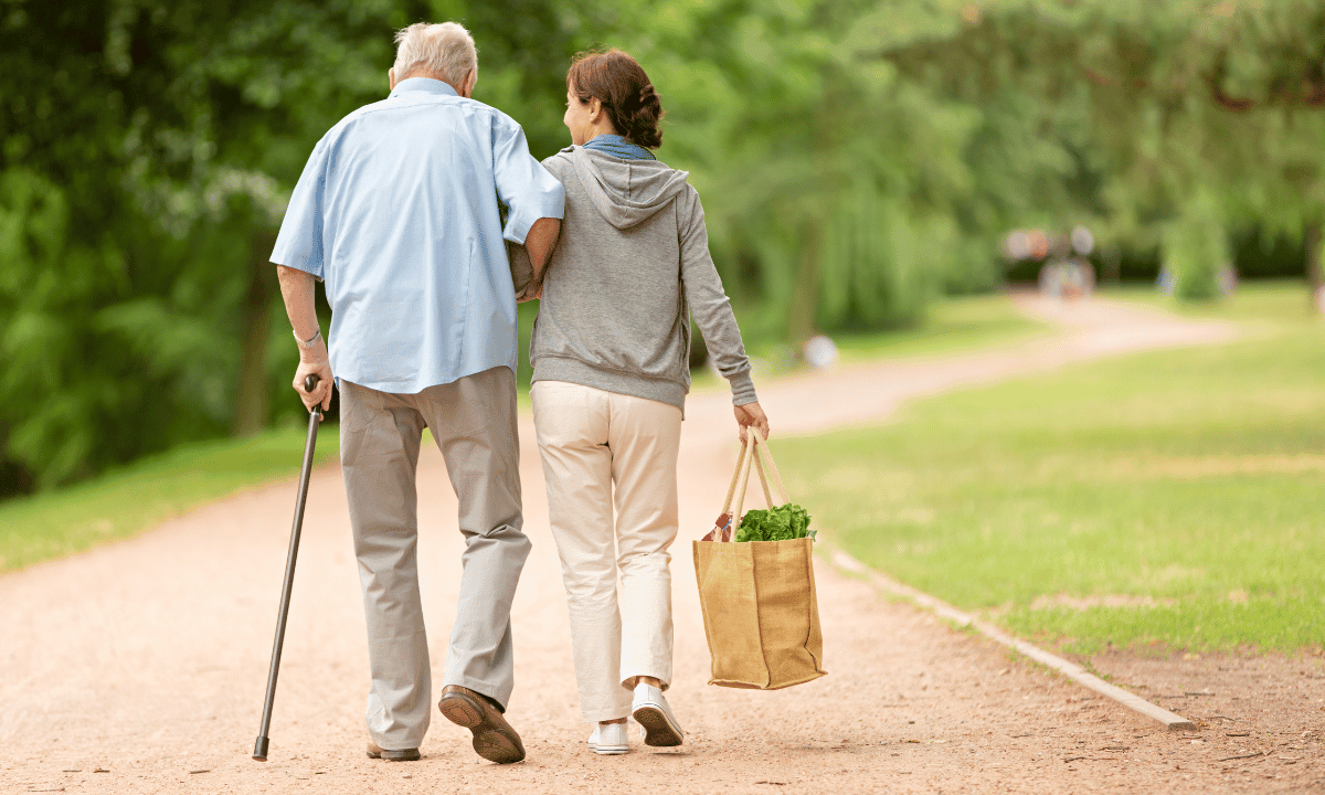 Un cuidador externo acompaña a un hombre mayor con bastón, caminando juntos por un parque.